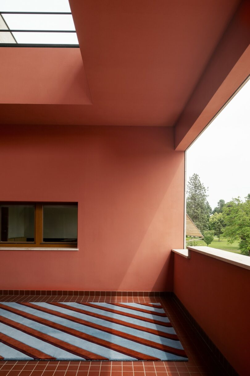 Una terraza exterior geométrica con paredes rojas, techo abierto, alfombra a rayas azules y rojas, ventanas con vistas a un paisaje de árboles verdes.