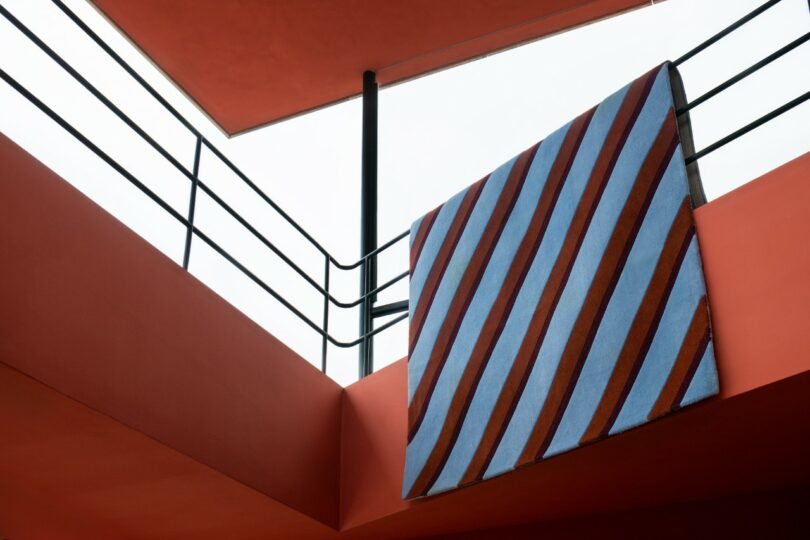 Una enorme pieza de tela de rayas diagonales azules y rojas con tiras de metal negro y cielo nublado colgando de una barandilla de balcón de color naranja brillante