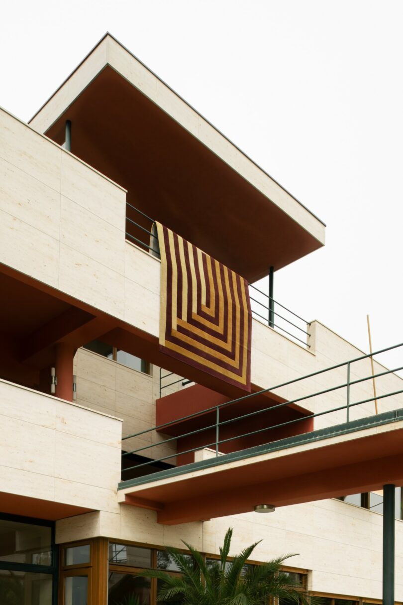 Una colcha con motivos geométricos marrones y amarillos cuelga de las barandillas de un moderno edificio de varios pisos con paredes beige y barandillas de metal oscuro