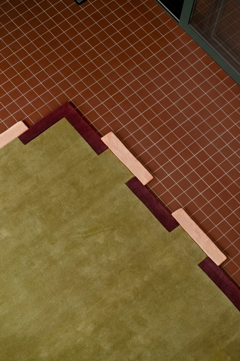 Alfombra verde geométrica con detalles en color burdeos y melocotón sobre un suelo de baldosas de color marrón rojizo cerca de una pared o puerta de cristal