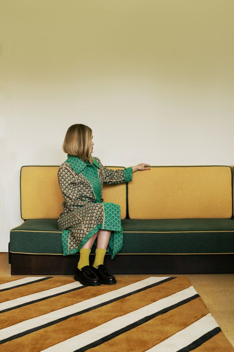 Una mujer rubia está sentada en un sofá verde y amarillo de estilo retro, con un abrigo estampado y calcetines amarillos, con los brazos apoyados en el respaldo del sofá.