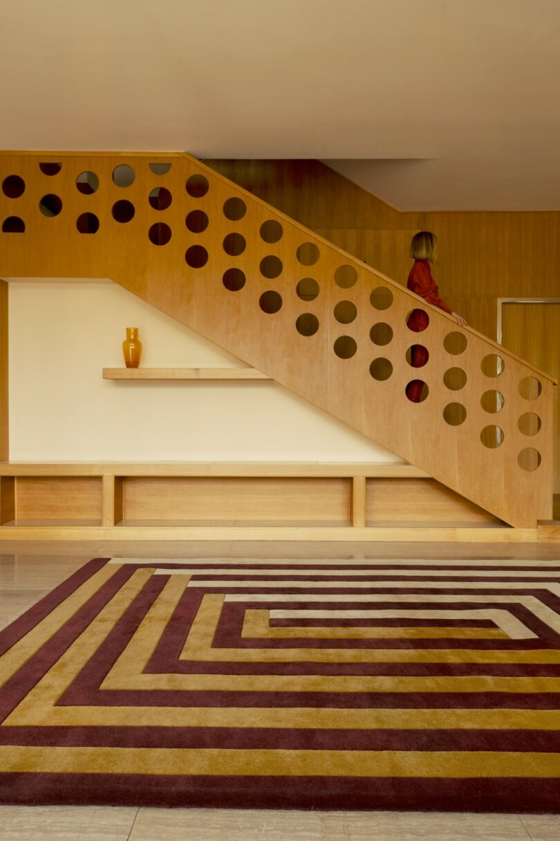 Una persona vestida de rojo sube una escalera de madera con un corte circular; En primer plano aparecen una alfombra con motivos geométricos y un jarrón sobre un estante.