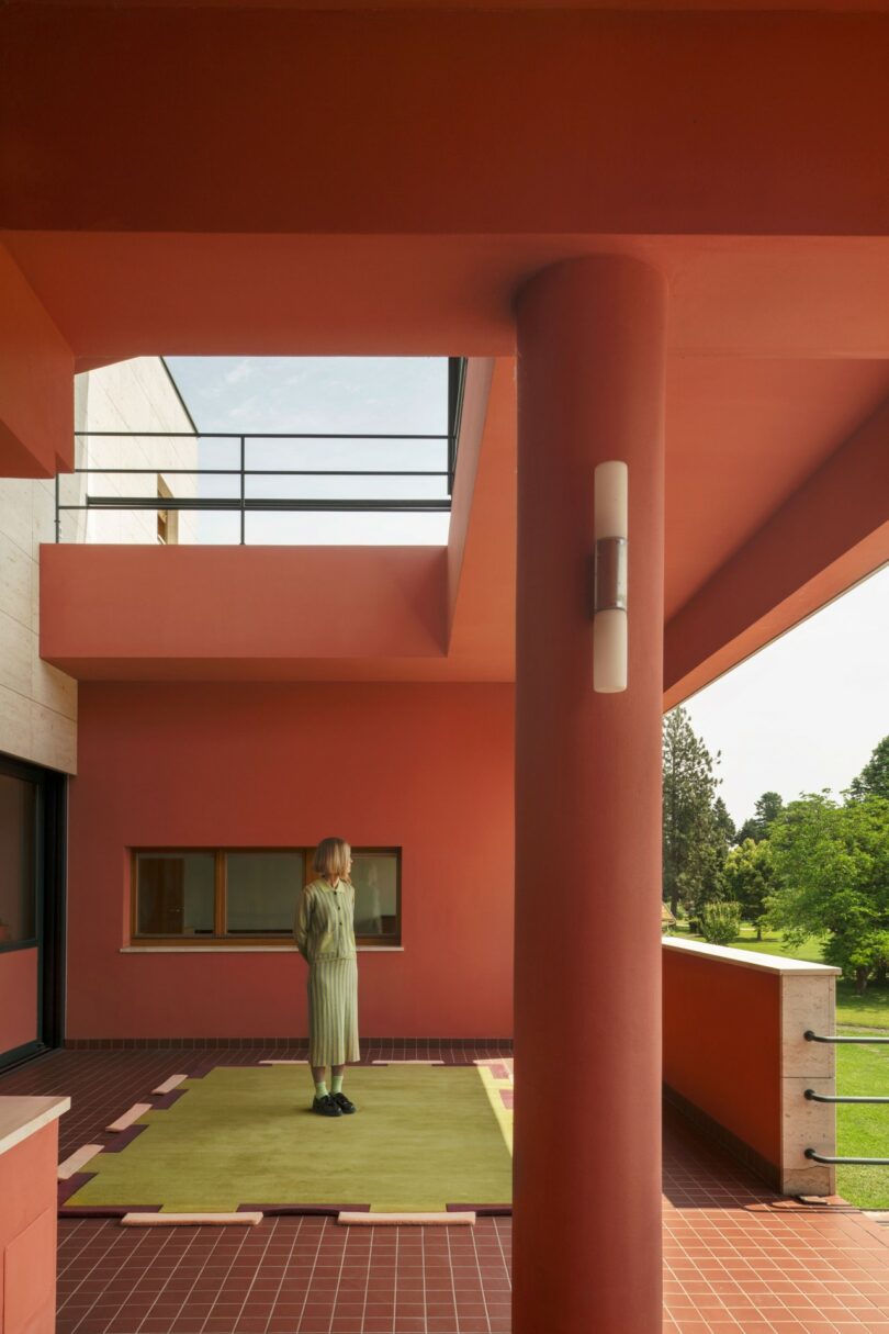 Una persona vestida de verde se alza sobre una alfombra amarilla en una terraza con azulejos rojos y columnas y paredes rojas, con vistas a un jardín verde