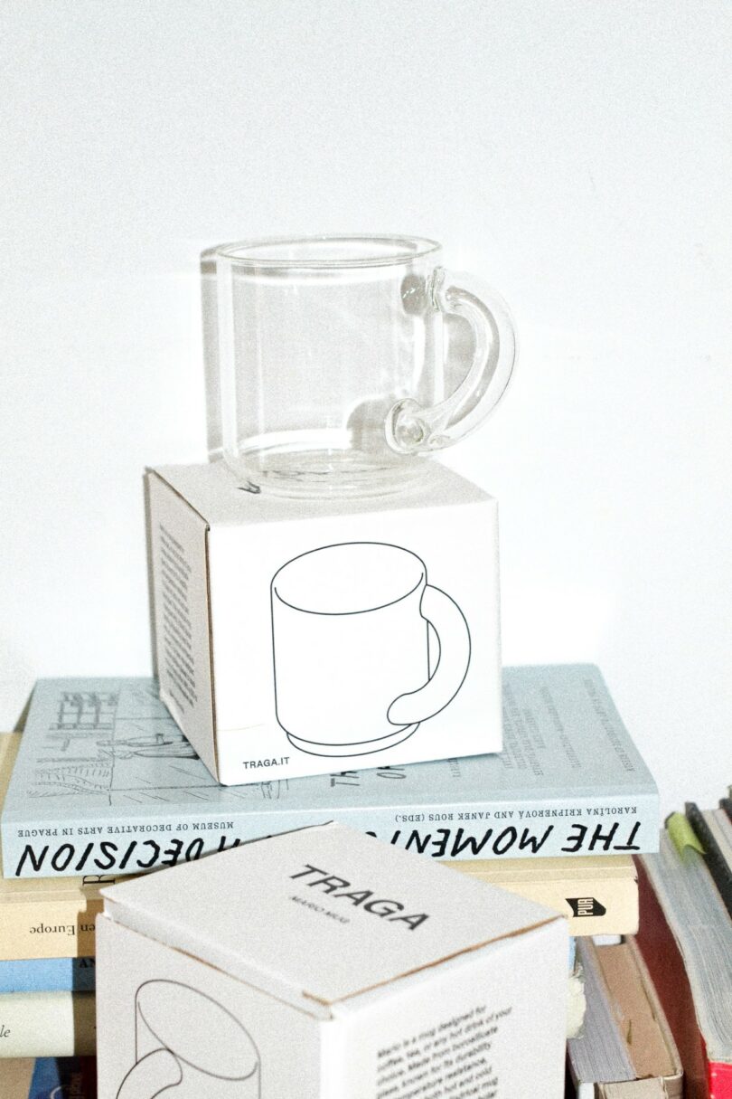 A clear glass mug sits on top of a white box with a mug illustration, surrounded by stacked books and another similar box in the foreground