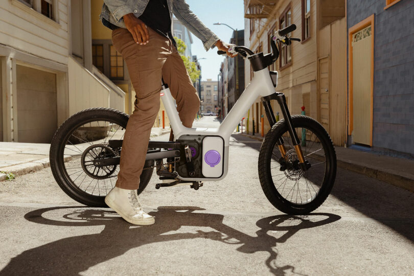 Person wearing light sneakers and brown pants stands next to a modern electric bike on a sunlit urban street.