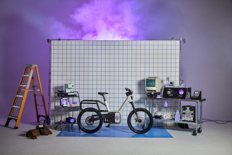 A white electric bicycle is displayed in front of a grid backdrop, surrounded by lab equipment, a yellow ladder, and purple smoke in the background.