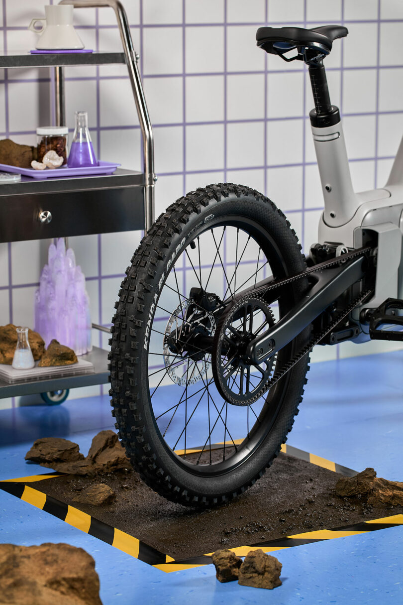 A close-up of a rugged electric bike’s rear wheel on a dirt patch, with rocks scattered around and lab equipment on a metal cart in the background.