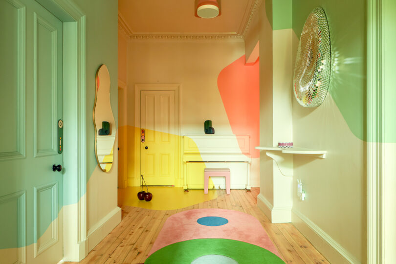 El pasillo tiene paredes y puertas de colores pastel, un piano amarillo, un espejo decorativo, alfombras de color rosa suave y verde y pisos de madera.