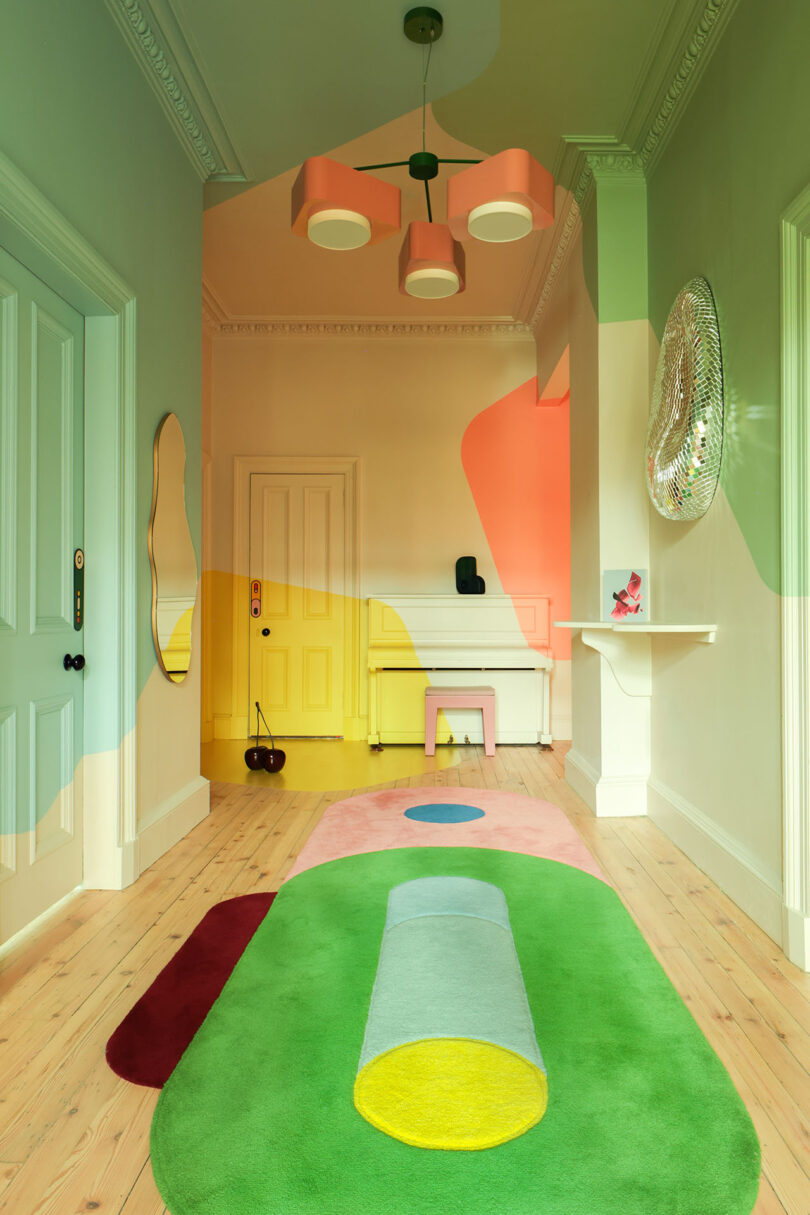 El pasillo presenta paredes en colores pastel, alfombras con motivos geométricos, lámparas modernas, puertas de color menta, detalles en amarillo y espejos de pared decorativos redondos.