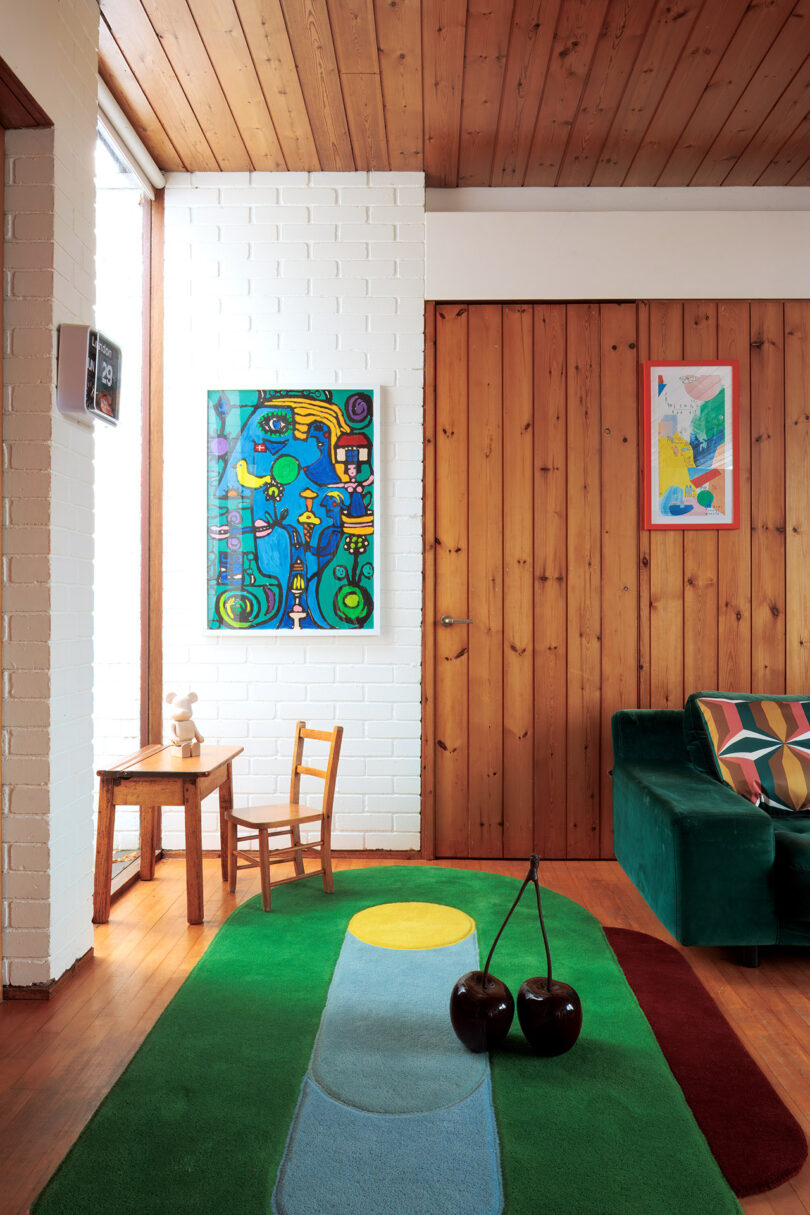 Salón con paredes de paneles de madera, sofá verde, alfombra colorida, pequeña mesa de madera con sillas, paredes de ladrillo blanco, dos obras de arte y un jarrón en el suelo.