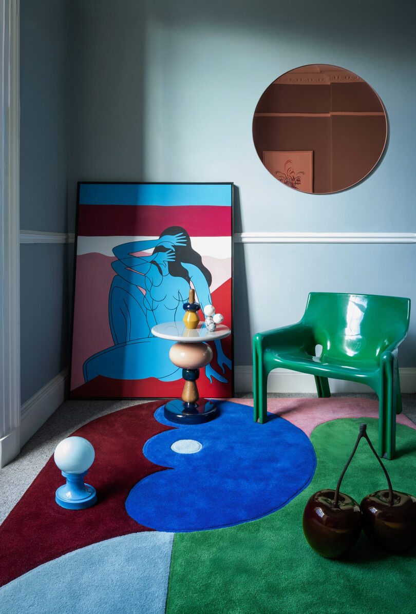 Una habitación moderna con una alfombra abstracta azul y roja, sillas verdes, un espejo redondo, coloridas láminas artísticas, una pequeña mesa con elementos decorativos y dos grandes esculturas de cerezos.