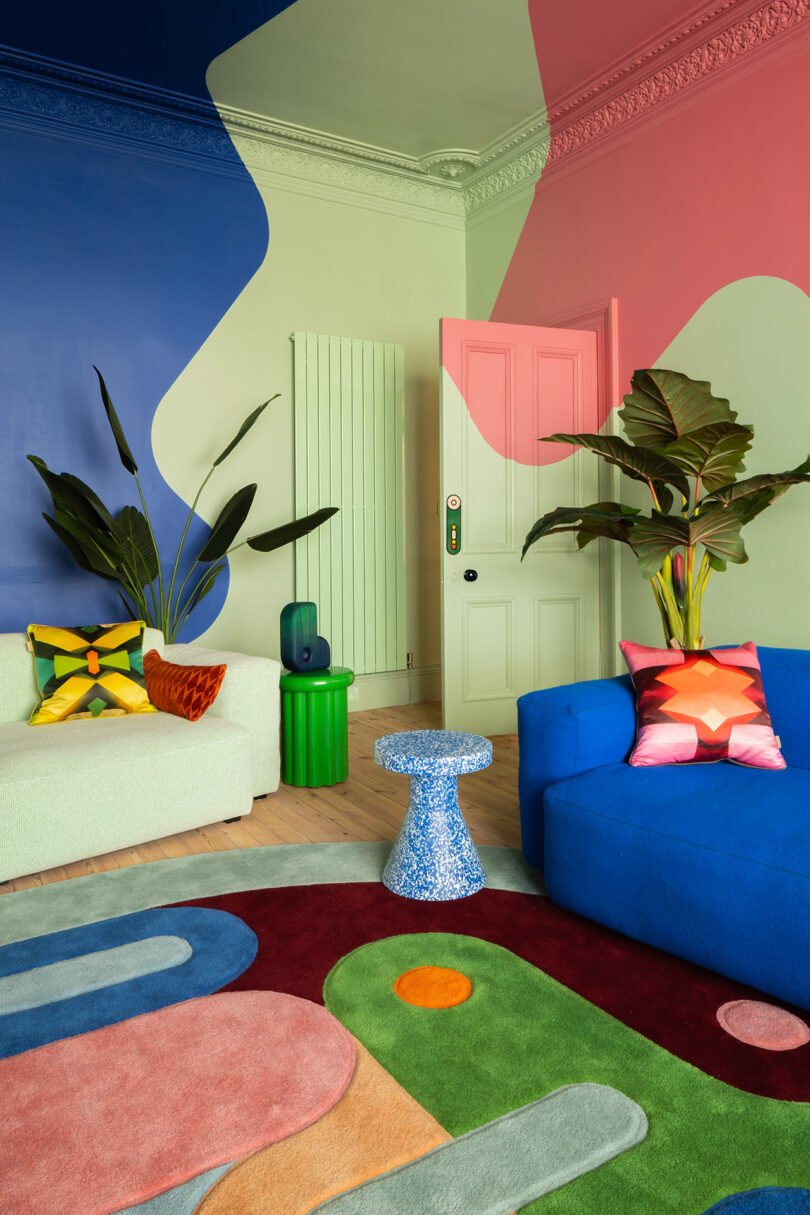 Salón colorido con paredes pintadas de forma abstracta, alfombra geométrica llamativa, sofá azul, sofá claro, plantas y almohadas estampadas.