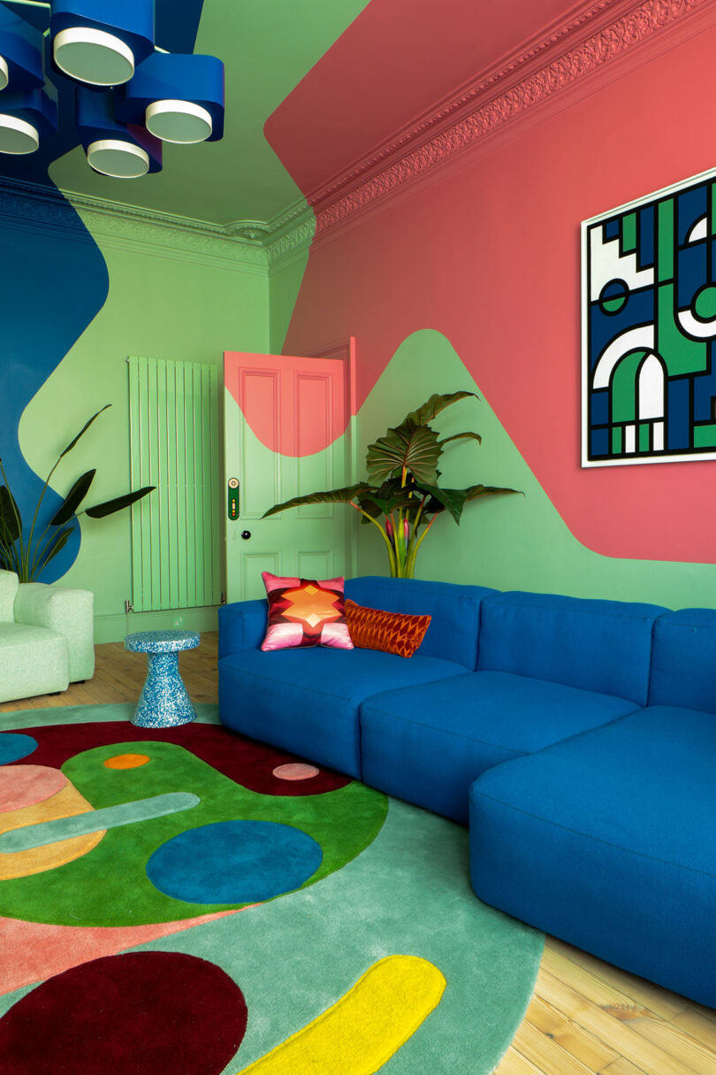 La colorida sala de estar presenta un atrevido patrón geométrico en las paredes, un sofá seccional azul, obras de arte gráficas, plantas y detalles coloridos.