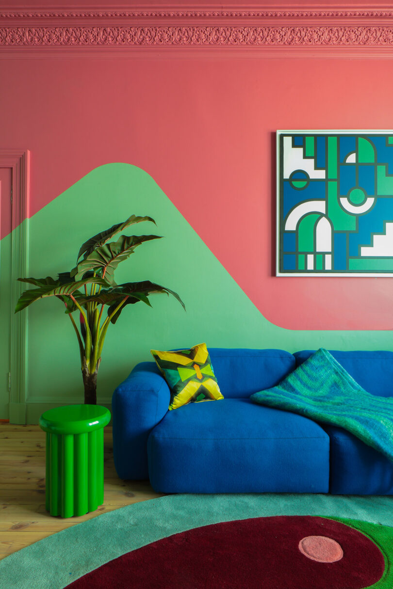 Salón con sofá azul, mesita verde, plantas, cojines geométricos, paredes verdes y rosas y arte abstracto colgado sobre el sofá.