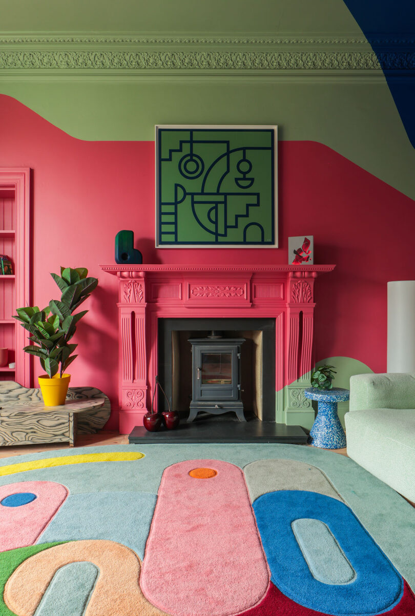 Sala de estar colorida con chimenea rosa, arte mural geométrico abstracto, paredes verdes, alfombras coloridas y muebles modernos con una maceta a la izquierda.