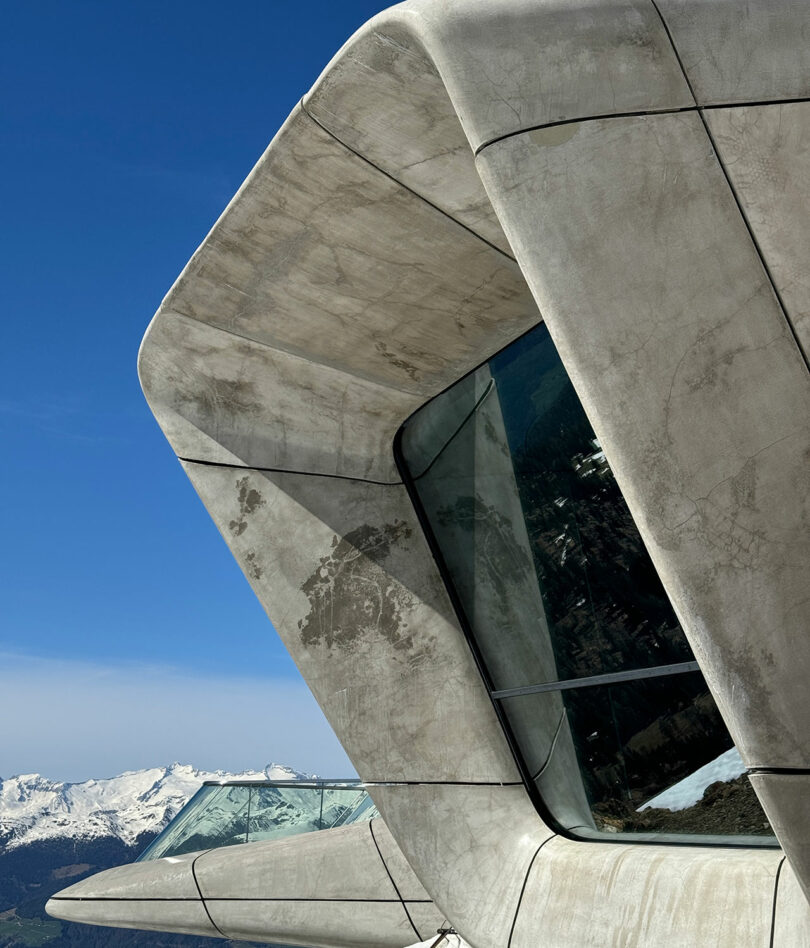 Vista cercana de un moderno edificio de hormigón en Camere con elementos angulares y grandes ventanales de cristal, con cielo azul y montañas nevadas al fondo.
