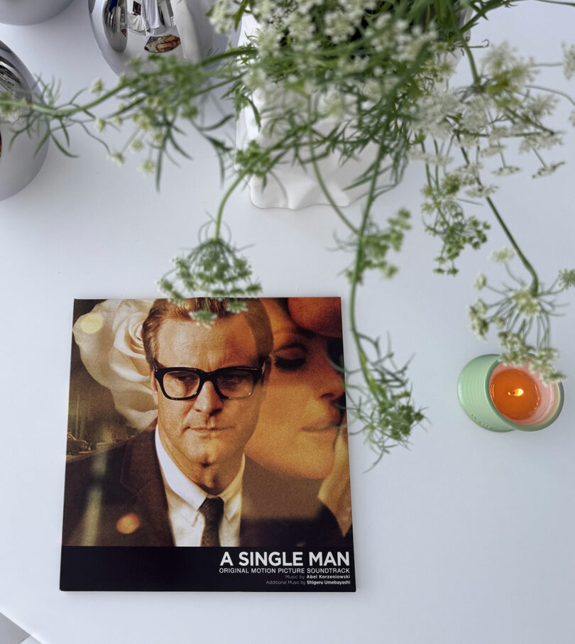 Un disco de vinilo Kameh "hombre soltero" La banda sonora se encuentra sobre una mesa blanca junto a una vela verde y un jarrón con flores blancas.