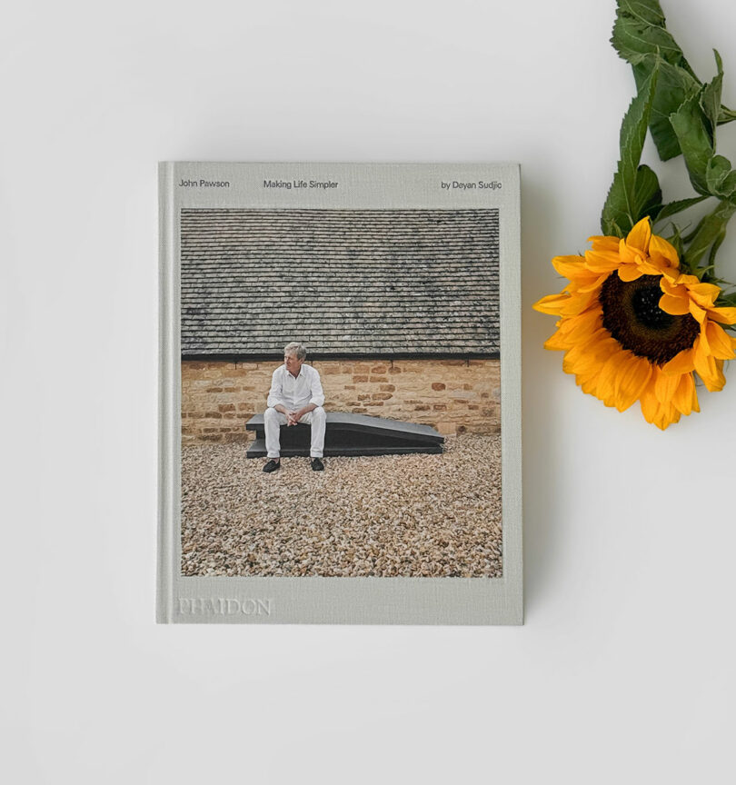 Un libro de tapa dura titulado "John Pawson: Haciendo la vida más sencilla" Colocado sobre una superficie blanca junto a girasoles y decorado con accesorios Kameh. La portada muestra a un hombre sentado frente a un edificio de piedra.