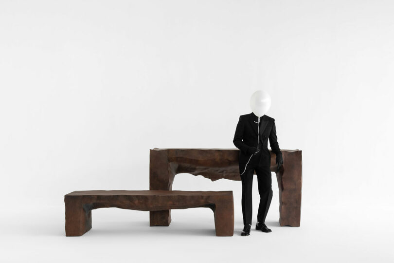 Un hombre con traje negro y un globo blanco en la cabeza se encuentra junto a dos bancos Kameh marrones de forma irregular sobre un fondo blanco puro.