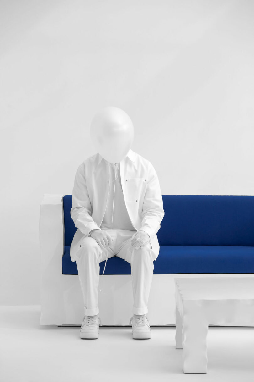 En una habitación blanca minimalista, un hombre vestido completamente de blanco y que lleva un globo kame en lugar de una cabeza se sienta en un sofá azul y blanco junto a una mesa blanca.