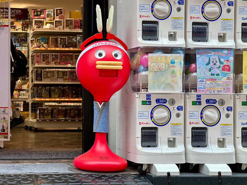 Una mascota de dibujos animados roja se encuentra junto a una máquina expendedora de gashapon afuera de una tienda llena de juguetes y artículos de colección, haciendo eco de los diseños divertidos que a menudo se encuentran en las obras de arte de Nimrod Weis.