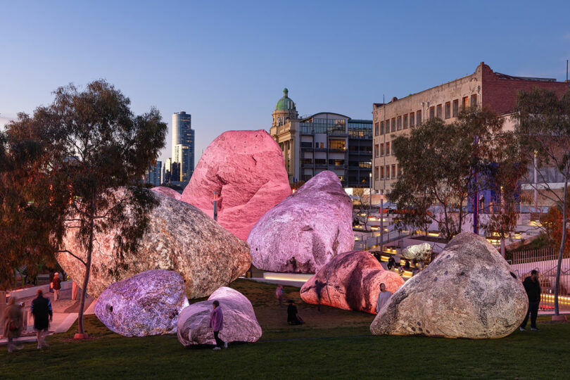 Grandes rocas artificiales diseñadas por Nimrod Weis se encuentran en un parque de la ciudad, iluminadas por luces rosas y violetas mientras la gente pasea cerca al anochecer.