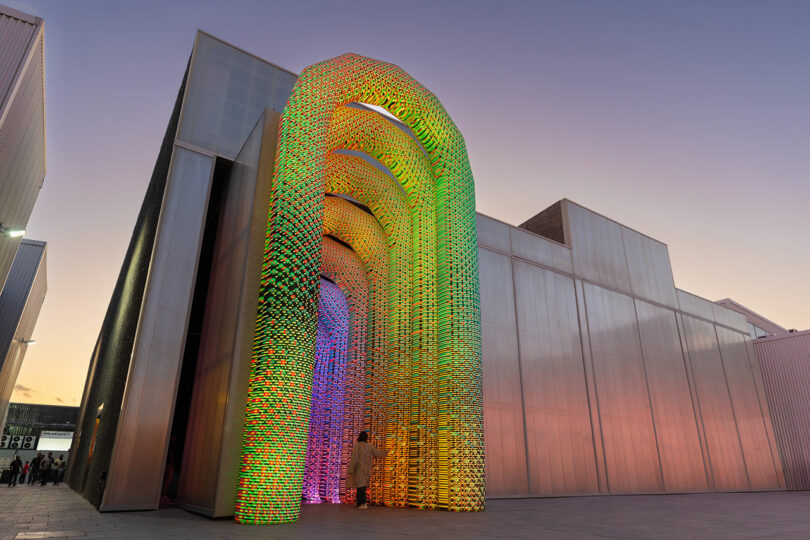 Una persona se encuentra en la base de un arco alto con los colores del arco iris diseñado por Nimrod Weis y hecho de materiales entrelazados afuera de un moderno edificio translúcido al atardecer.