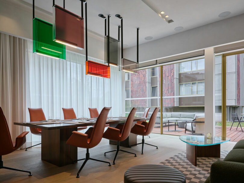 Moderna sala de conferencias con sillas marrones, una gran mesa de madera, coloridas lámparas rectangulares y ventanales con vistas al balcón y a la arquitectura contemporánea.