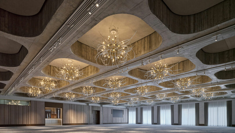 El espacioso y moderno salón de baile cuenta con grandes ventanas, un techo con un diseño único y múltiples candelabros modernos distribuidos uniformemente por toda la sala.