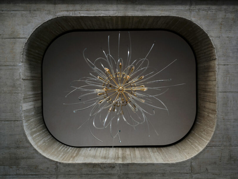 Una moderna lámpara de araña escultural con elementos dorados y de cristal transparente cuelga del techo, que cuenta con una gran abertura rectangular redondeada de hormigón.