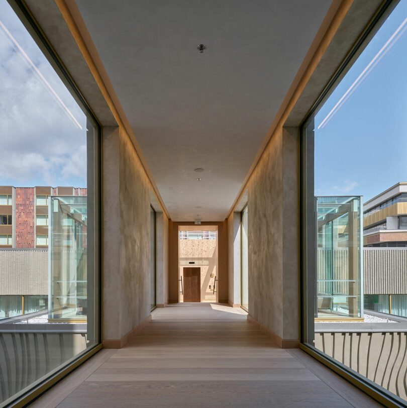 Un moderno pasillo con paredes de vidrio, pisos de madera y paredes de color beige ofrece vistas de los edificios vecinos y del cielo azul del exterior.