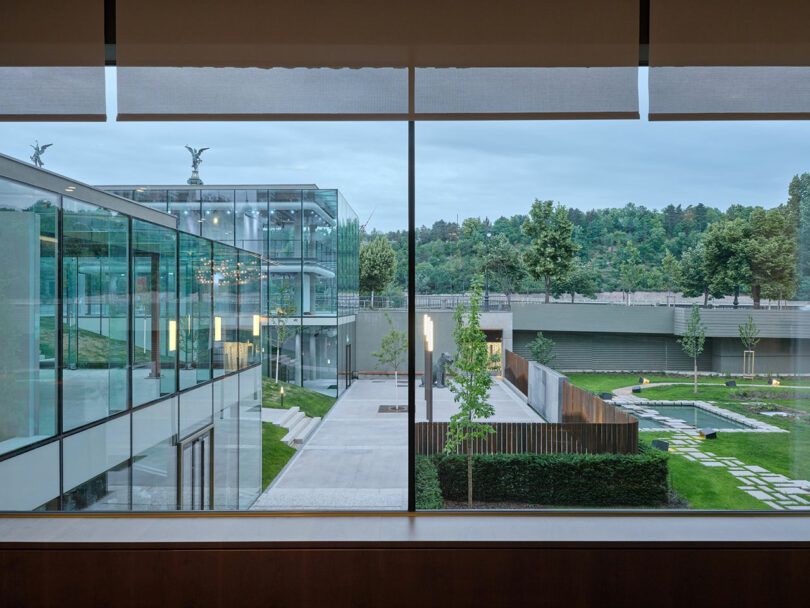 Un gran ventanal revela modernos edificios de cristal, jardines, senderos, árboles y espacios verdes bajo un cielo nublado.