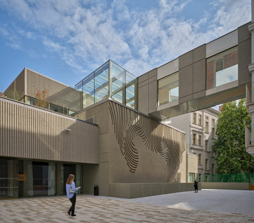 Edificio moderno con fachada texturizada, puente de cristal y arte mural geométrico; una persona camina por un patio pavimentado bajo un cielo parcialmente nublado.