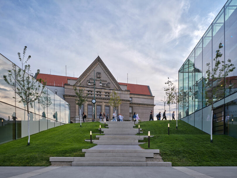 Un grupo de personas subía y bajaba las escaleras cubiertas de hierba que conducían a un edificio histórico con un reloj, rodeado por una moderna estructura de vidrio.