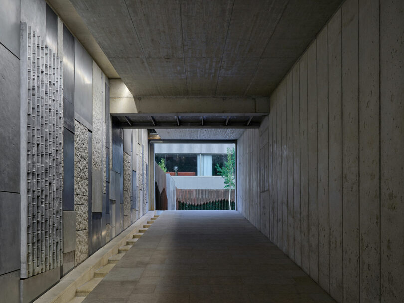 A la izquierda, un corredor de hormigón con paneles de pared texturizados conduce a un área abierta con árboles y un edificio moderno visible al fondo.
