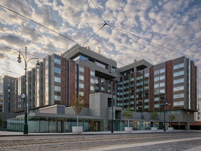 Moderno edificio de varios pisos con fachada de vidrio y metal, farola, reloj, cables de tranvía y calle adoquinada bajo un cielo parcialmente nublado en primer plano.