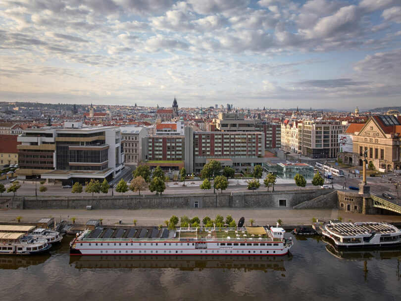 Un paisaje urbano con barcos fluviales atracados en primer plano, edificios modernos e históricos a lo lejos y un cielo parcialmente nublado.