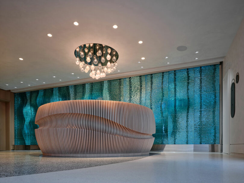 El moderno vestíbulo cuenta con un escultural mostrador de recepción de madera, paredes de vidrio azul texturizado y lámparas de techo con bombillas de vidrio colgantes.