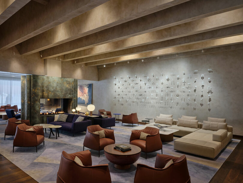 Salón moderno con sillas de cuero marrón, sofá beige, chimenea central, mesa baja e instalación de arte en las paredes texturizadas bajo vigas expuestas en el techo.