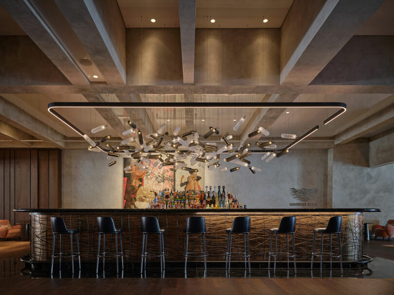 El moderno bar cuenta con un techo alto con una lámpara abstracta, seis taburetes negros, botellas alineadas detrás del mostrador y un gran mural en la pared trasera.