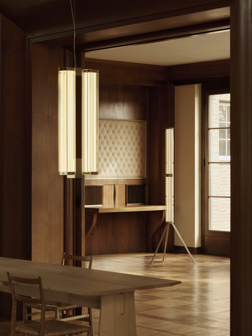 La habitación minimalista cuenta con paredes con paneles de madera, un escritorio incorporado con un panel trasero acolchado, modernas lámparas verticales y grandes ventanas que dejan entrar luz natural.