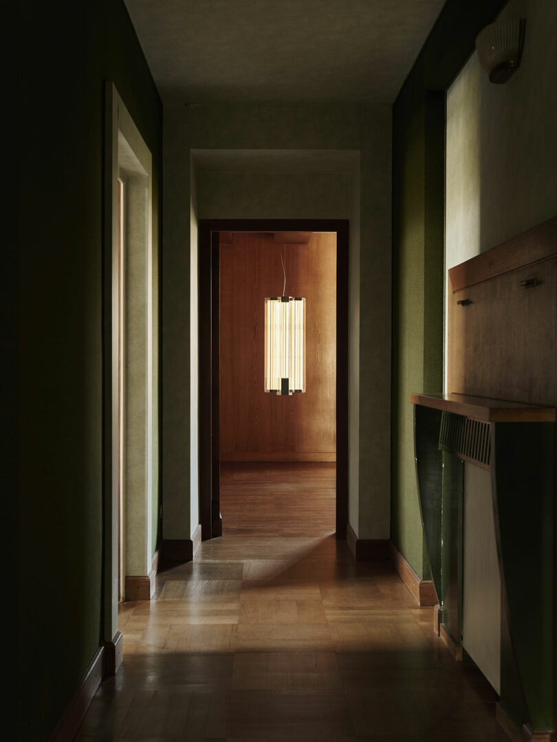 Un pasillo poco iluminado con paredes verdes conduce a una habitación de madera con una moderna lámpara de araña colgando del techo.