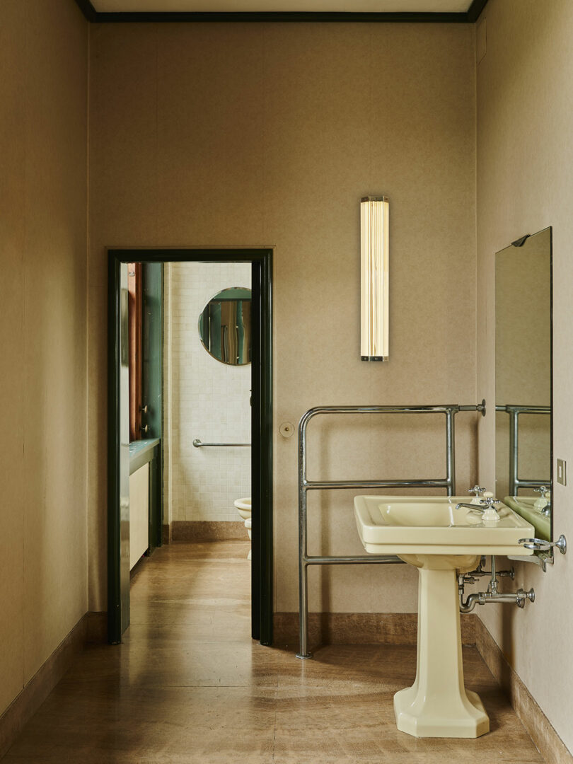 El baño beige cuenta con un lavabo con pedestal, un espejo de pared, un toallero de metal y una lámpara vertical; la puerta se abre para revelar un segundo baño con espejo redondo e inodoro.