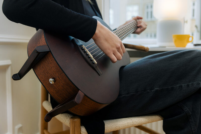 Person sitting on a chair indoors, playing a wooden acoustic guitar with a yellow cup on the table in the background.