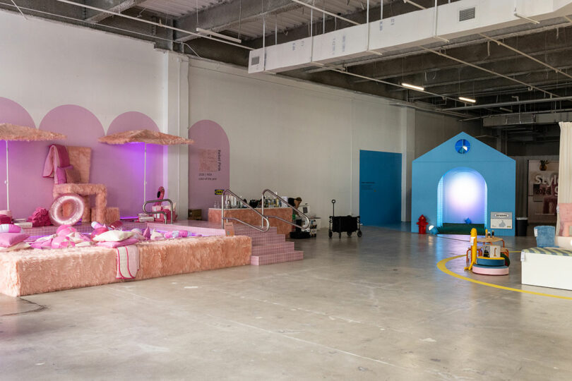 An interior space features playful installations in pastel colors, including a pink living room and a blue house structure, with exposed industrial ceilings above.