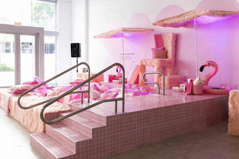 A pink-themed indoor display features faux fur furniture, pink tiled steps, umbrellas, throw pillows and a large inflatable flamingo with purple lighting.