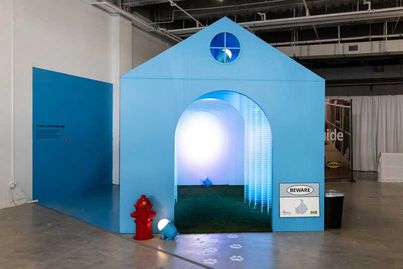 Large blue dog house construction with arched entrance, lit interior, artificial grass, red fire hydrant and "Watch" sign, displayed indoors on a concrete floor.
