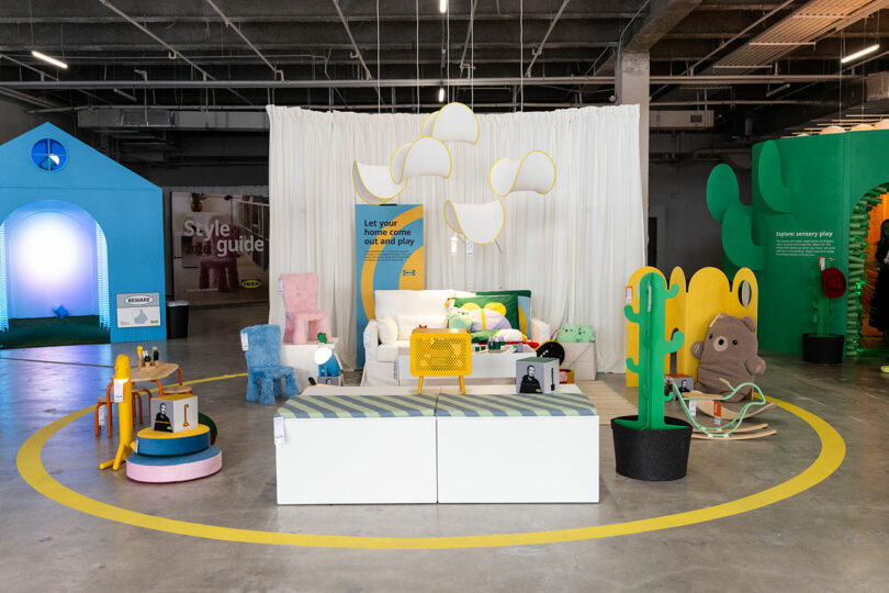 A colorful showroom showcases playful, modern furniture and decor with odd shapes and bright colors, on a concrete floor with a white curtain background and bright lighting.