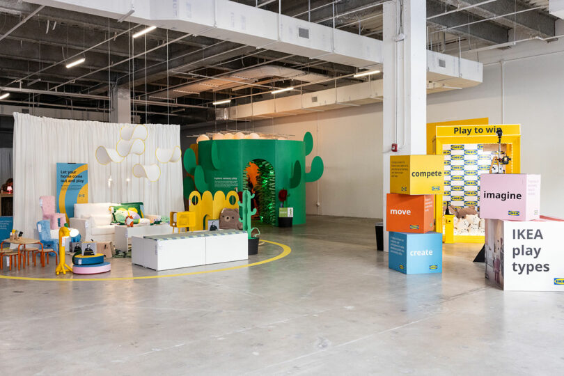 An indoor IKEA showroom features colorful furniture displays, playful decor, large signage and interactive zones in a spacious industrial-style environment.