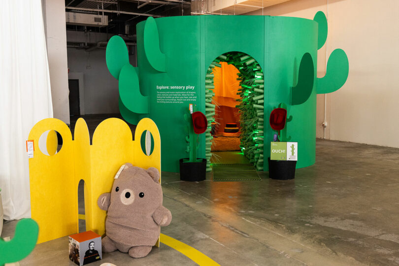 Green cactus themed sensory play facility with arched entrance, yellow abstract partition, teddy bear toy and concrete floors indoors.